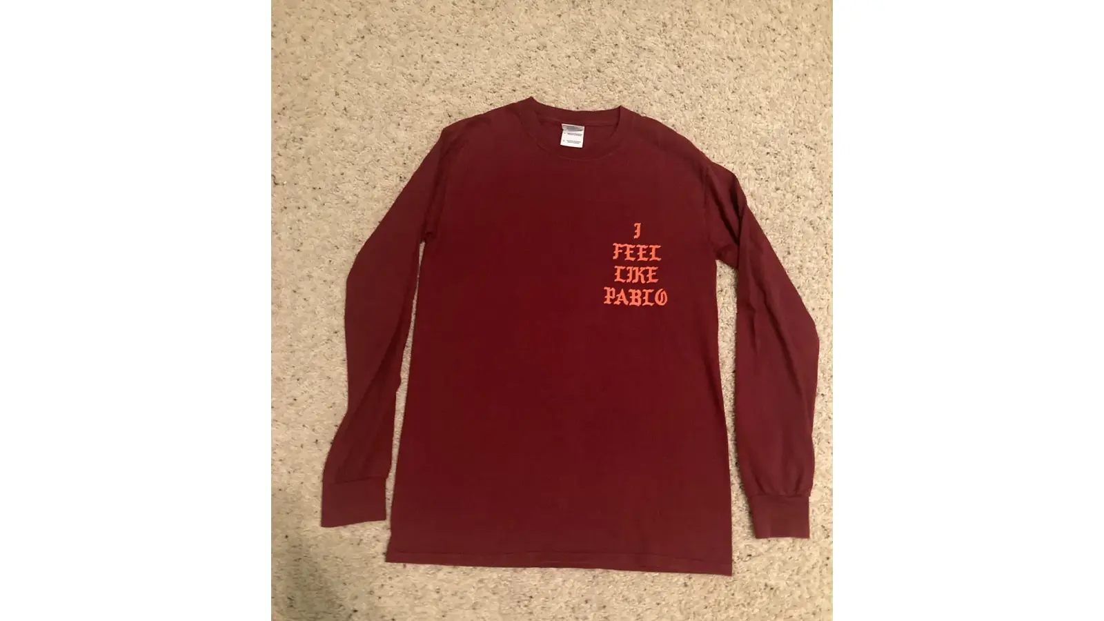 A flat-lay view of the "I Feel Like Pablo" merch longsleeve from the 2016 pop-up shops. The image focuses on the blue gothic lettering by Cali DeWitt against the vibrant orange fabric, showcasing the industrial-print aesthetic.