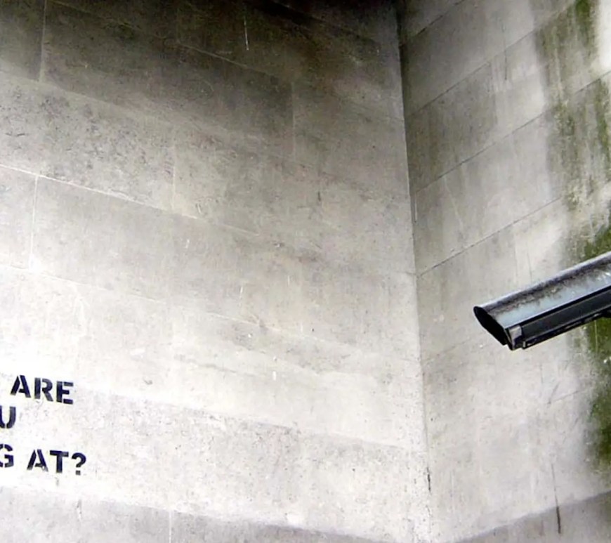 A sleek, high-tech security camera mounted on a city wall, symbolizing the invisible data collection and algorithmic surveillance of urban communities.