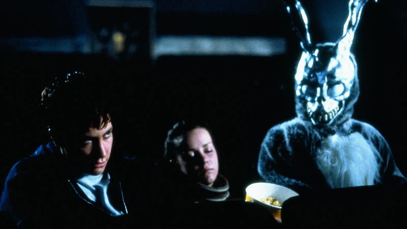 A high-definition 4K restoration still from Donnie Darko (2001) featuring Jake Gyllenhaal in a high school hallway as a CGI "liquid spear" vector emerges from his chest. The image showcases the heavy, atmospheric grain of the Kodak Vision 800T film stock and the characteristic soft blue "bloom" created by Panaflex Primo Anamorphic lenses and Pro-Mist filters.
