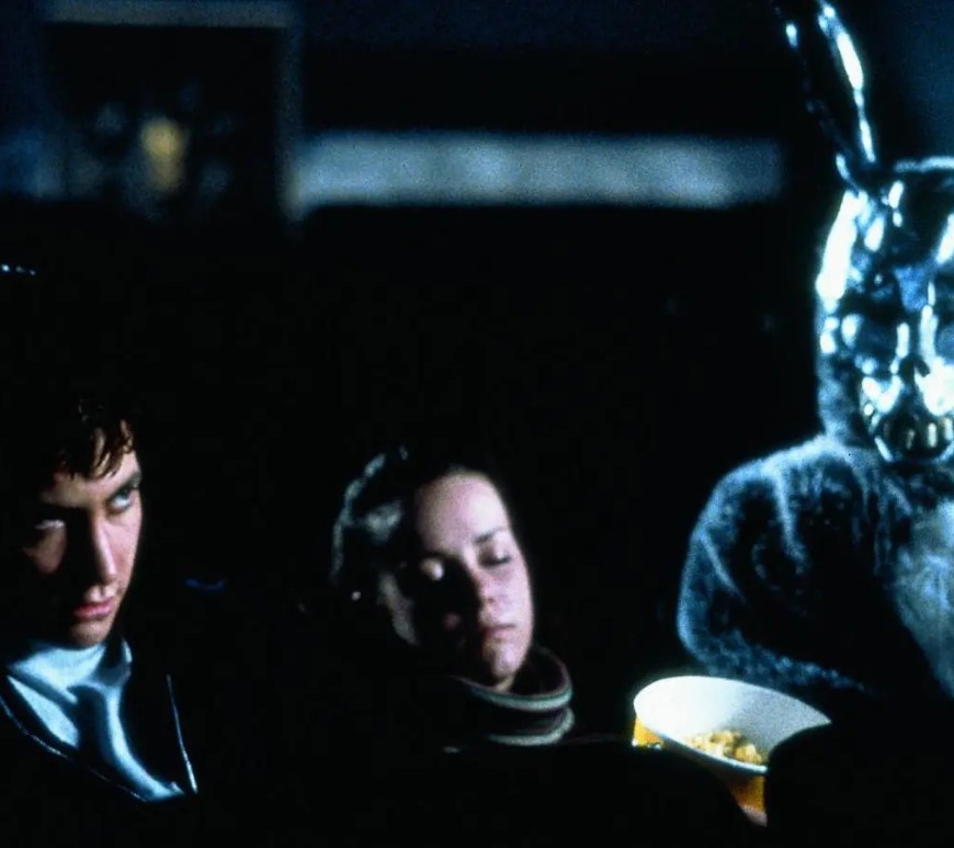 A high-definition 4K restoration still from Donnie Darko (2001) featuring Jake Gyllenhaal in a high school hallway as a CGI "liquid spear" vector emerges from his chest. The image showcases the heavy, atmospheric grain of the Kodak Vision 800T film stock and the characteristic soft blue "bloom" created by Panaflex Primo Anamorphic lenses and Pro-Mist filters.