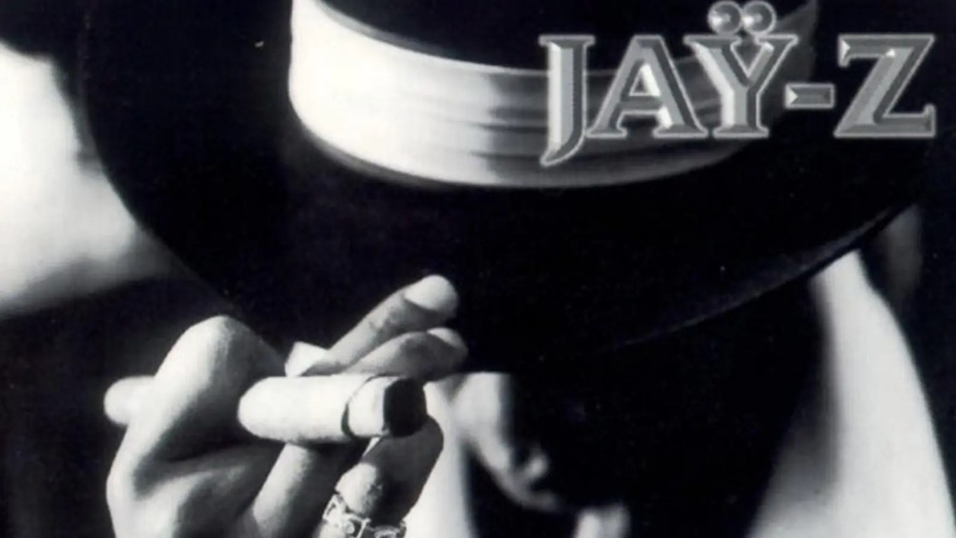 The black-and-white cover art for Jay-Z's 1996 debut album Reasonable Doubt, featuring Shawn Carter in a classic 1920s-style Fedora, wool overcoat, and a white scarf, holding a cigar—shot by photographer Jonathan Mannion.