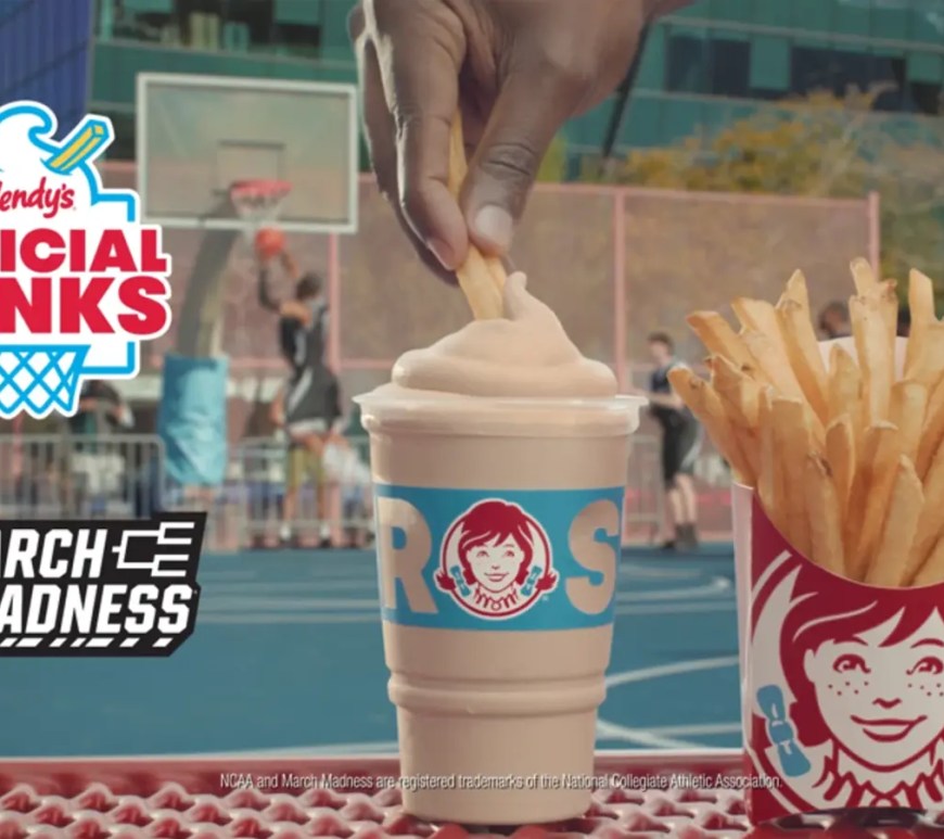 A high-resolution screenshot of the Wendy’s mobile app interface featuring the 2026 March Madness 'Dunks Menu.' The design uses a dark-mode aesthetic with vibrant orange and blue accents, showcasing the 'Starting Lineup' of a Classic Frosty and Hot & Crispy Fries. Promotional text highlights the 'Dunkstakes' entry for app-based purchases and a teaser for the Phase 2 expansion including Nuggets and Tenders.