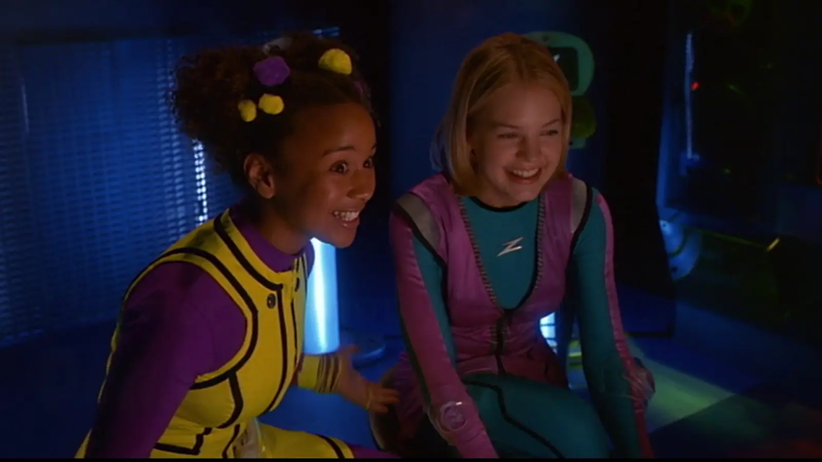 Kirsten Storms as Zenon Kar and Shadia Simmons as Nebula Wade in their metallic 2001 'Zequel' outfits, looking at a Zap-Pad in the Alien Patrol lab.