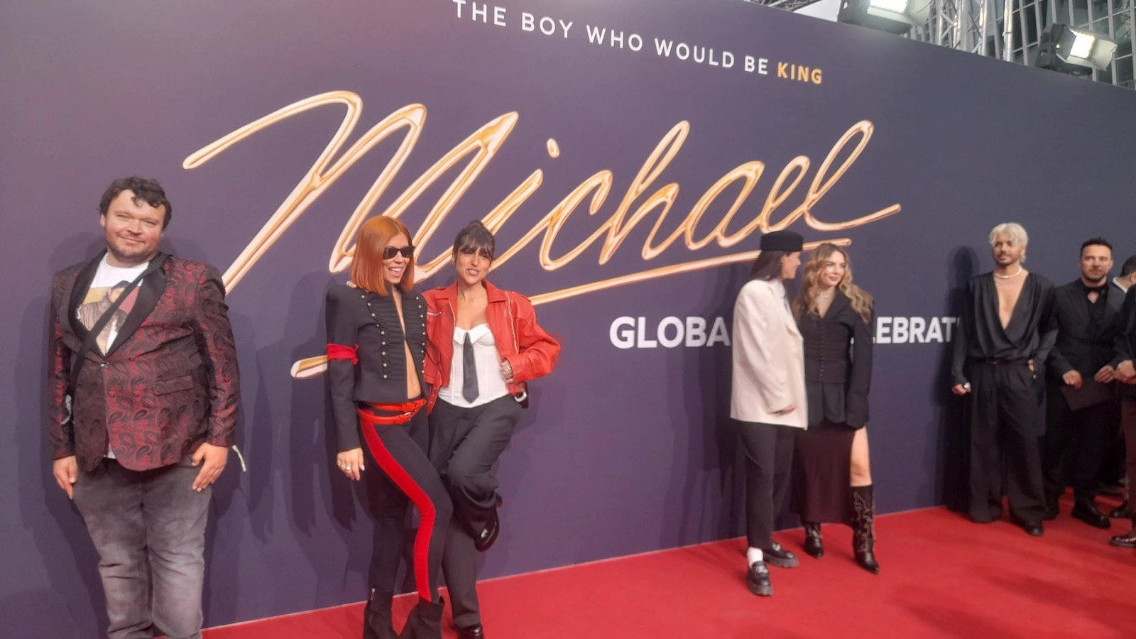 Michael Movie World Premiere Berlin: A group of attendees stands on a red carpet in front of a large premiere backdrop reading “The Boy Who Would Be King” and “Michael” in gold script at the world premiere of Michael in Berlin, Germany. On the far left, Decked Out Magazine correspondent Giorgio Tardio poses confidently in front of the step‑and‑repeat, dressed in event attire, with other guests lined up beside him against the branded wall.