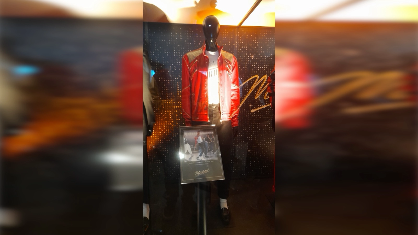 MICHAEL: The Greatest Musical Biopic of All Time: Michael Movie World Premiere Berlin: Mannequin displaying Jaafar Jackson’s red leather ‘Beat It’ costume from the film MICHAEL, featuring a red zip‑front jacket, white T‑shirt, black pants, white socks, and black shoes, shown inside a glass case with a photo of the matching performance outfit beneath it.