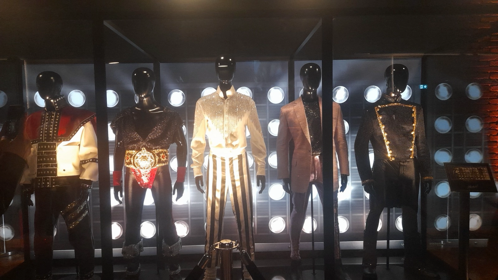 The Greatest Musical Biopic of all time: Display of five mannequins dressed in elaborate stage costumes from the film MICHAEL at the Michael Movie World Premiere Berlin, including sequined jackets, metallic fabrics, embellished shirts, and performance belts, arranged in front of a wall of circular stage lights to evoke a concert‑style atmosphere.