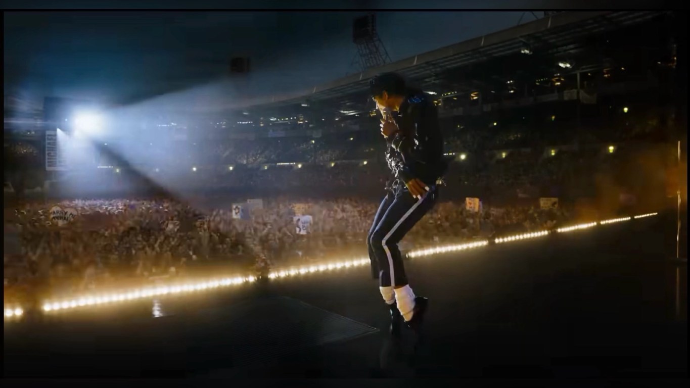 MICHAEL: The Greatest Musical Biopic of All Time: Jaafar Jackson performing as Michael Jackson on a stadium stage, captured mid‑dance in a black jacket, dark pants with white stripes, white socks, and black shoes, illuminated by a strong spotlight as a massive cheering crowd fills the arena.