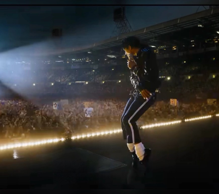 MICHAEL: The Greatest Musical Biopic of All Time: Jaafar Jackson performing as Michael Jackson on a stadium stage, captured mid‑dance in a black jacket, dark pants with white stripes, white socks, and black shoes, illuminated by a strong spotlight as a massive cheering crowd fills the arena.