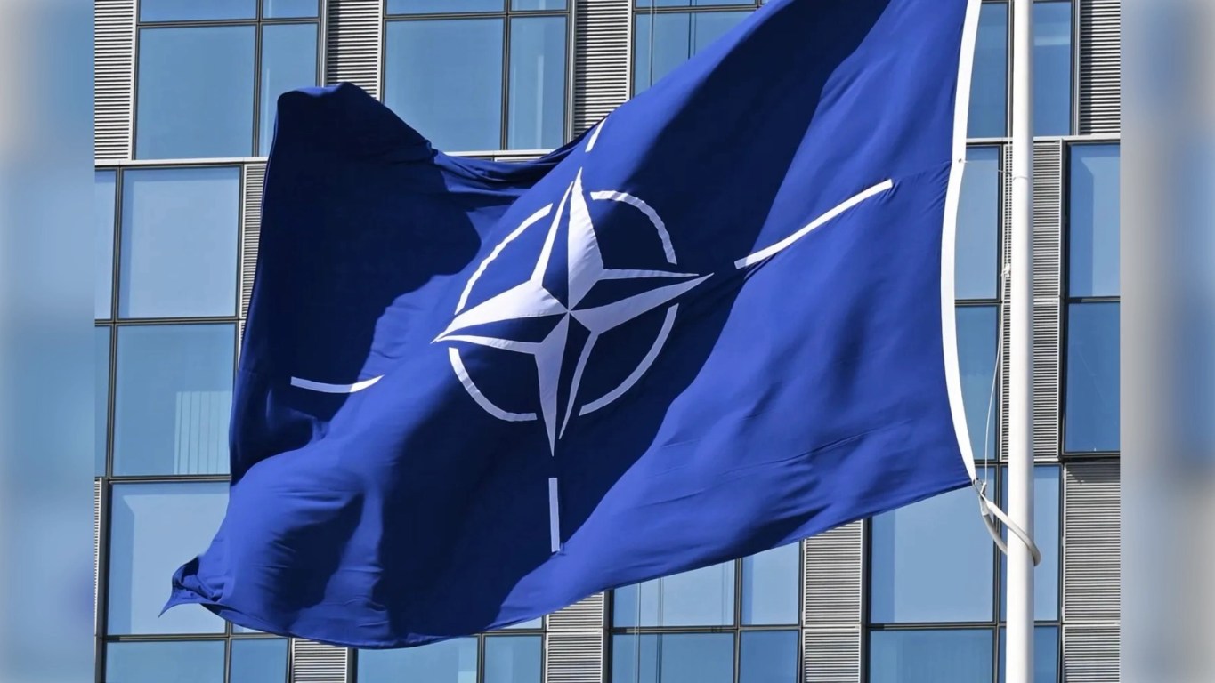 the United States is More Isolated Than Ever Before: NATO flag flying in front of the glass façade of NATO headquarters, showing the white compass rose emblem against a deep blue field.