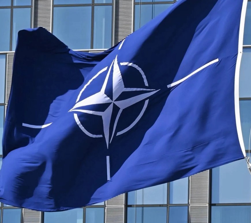 the United States is More Isolated Than Ever Before: NATO flag flying in front of the glass façade of NATO headquarters, showing the white compass rose emblem against a deep blue field.