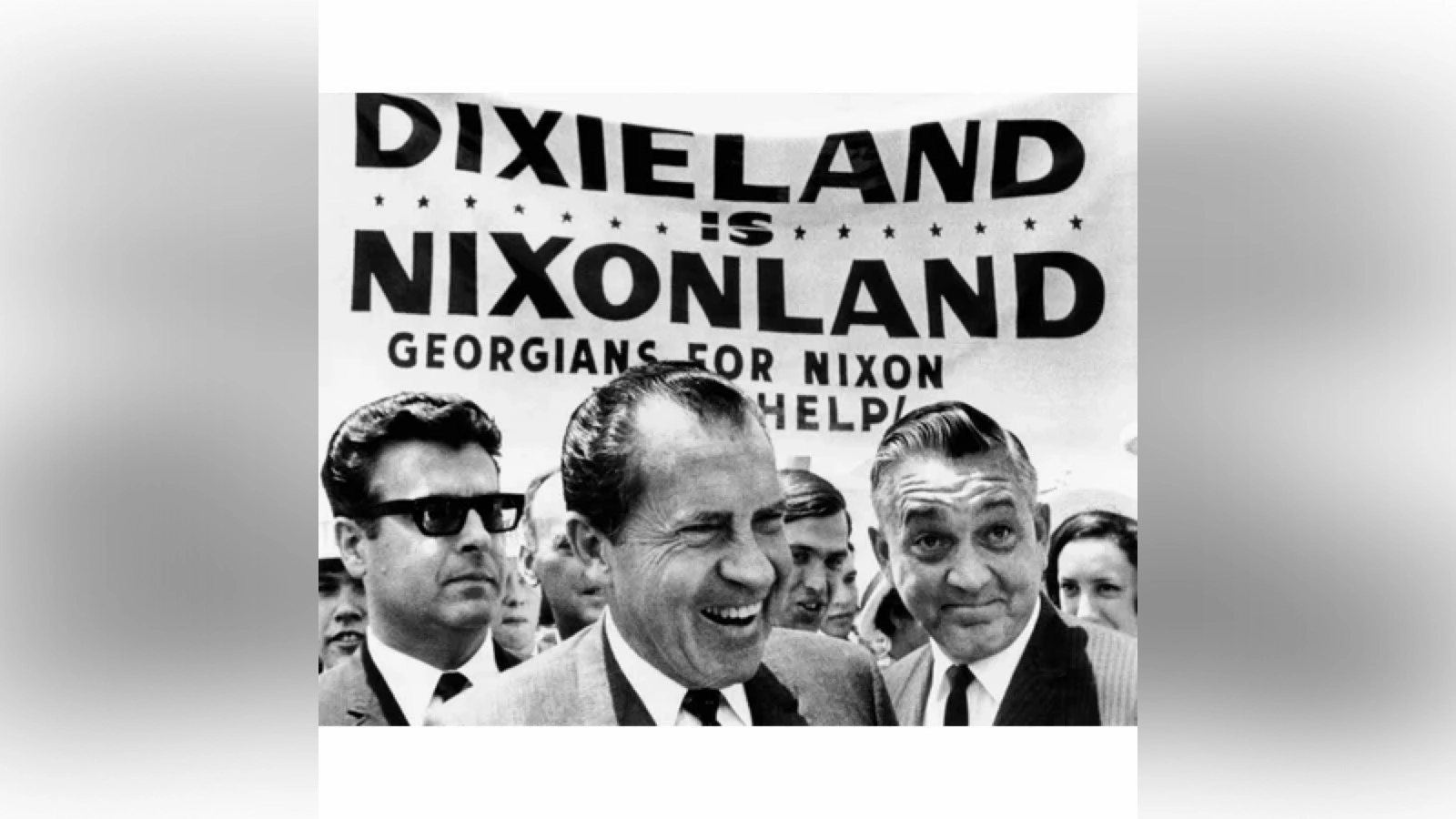 
'Democrats are the party of the KKK' is a Falsehood: Black‑and‑white photo of Richard Nixon standing with supporters in front of a large campaign banner reading ‘Dixieland Is Nixonland — Georgians for Nixon Help!,’ reflecting his appeal to white Southern voters during the 1968 campaign.