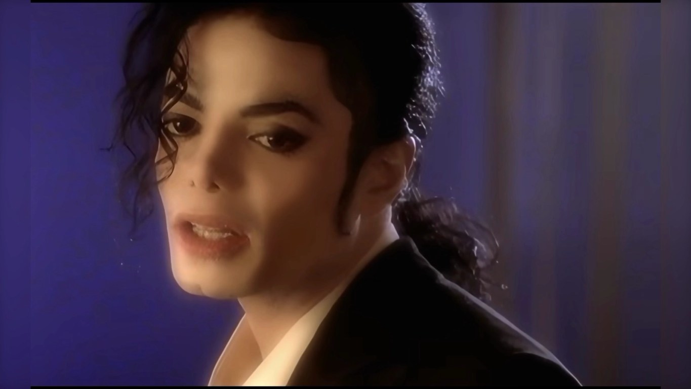 Is Who is It Michael Jackson's greatest deep cut? Close‑up still from a music short film showing a person with styled dark hair and a single curl over the forehead, wearing a dark jacket and white shirt, lit dramatically against a blue‑purple background.