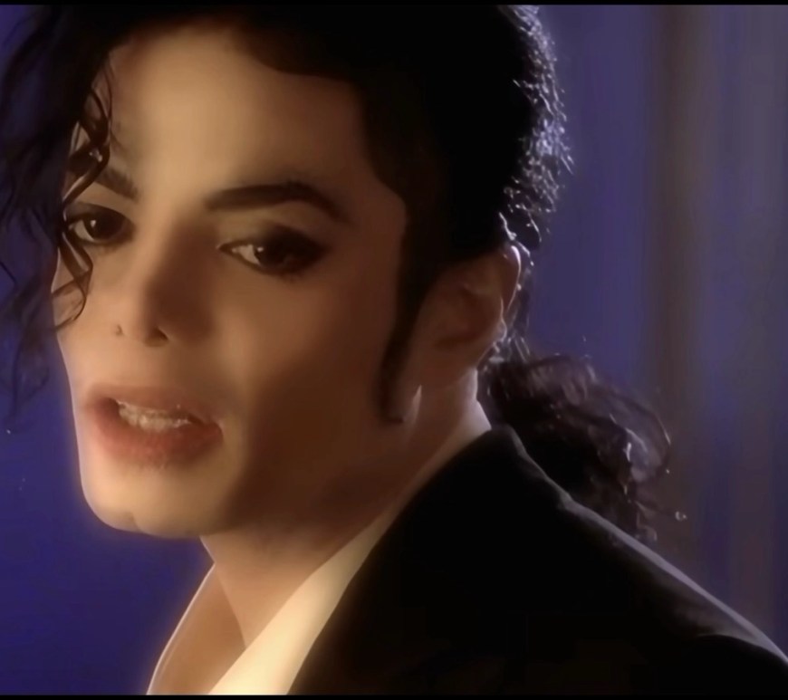 Is Who is It Michael Jackson's greatest deep cut? Close‑up still from a music short film showing a person with styled dark hair and a single curl over the forehead, wearing a dark jacket and white shirt, lit dramatically against a blue‑purple background.