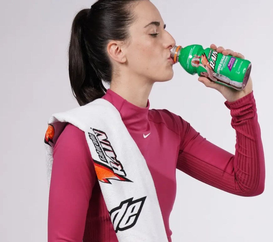 A promotional portrait of Caitlin Clark for the 2026 Gatorade 'Rain Berry' relaunch. Clark stands confidently in her white and gold Indiana Fever practice gear, holding a commemorative 20 oz. Gatorade bottle.