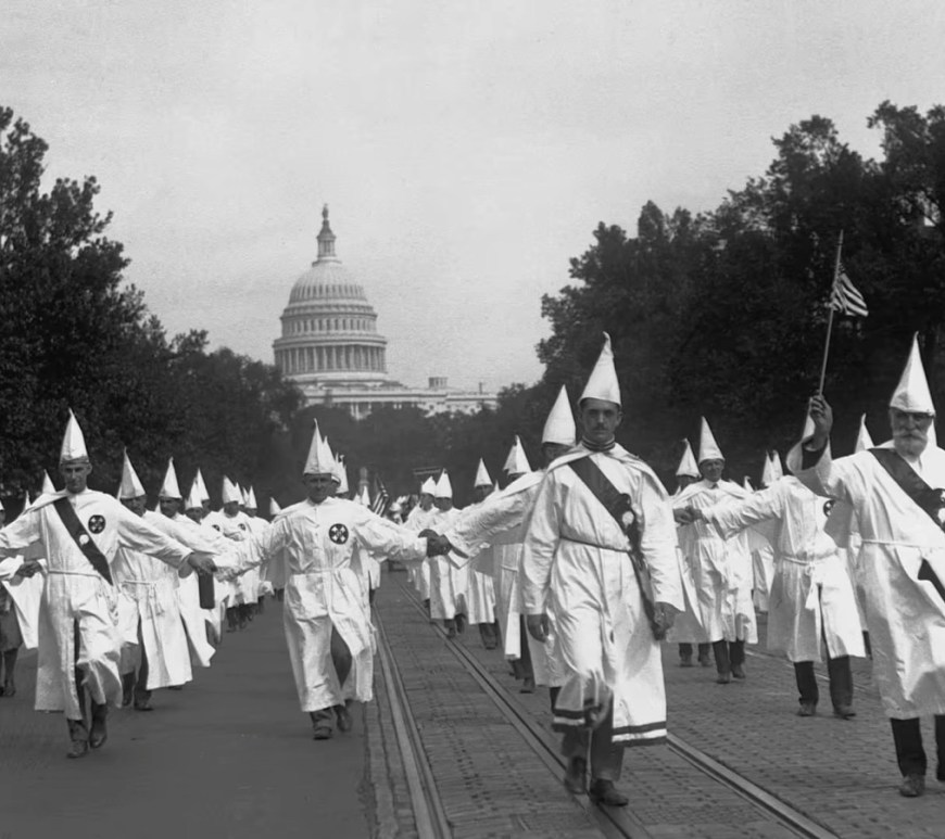 'Democrats are the party of the KKK' is a Falsehood: Black‑and‑white photograph of Ku Klux Klan members in white robes and pointed hoods marching in formation down a Washington, D.C. street with the U.S. Capitol visible in the background.