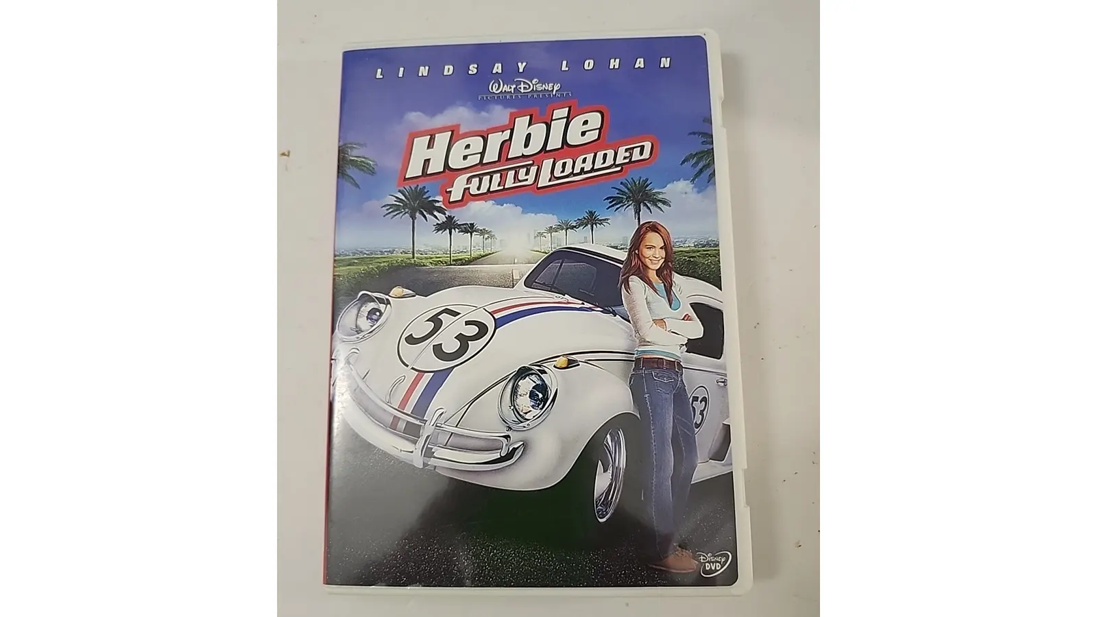 Lindsay Lohan standing with the 1963 NASCAR-spec Herbie Volkswagen Beetle on a race track, featuring wider tires and a custom roll cage.