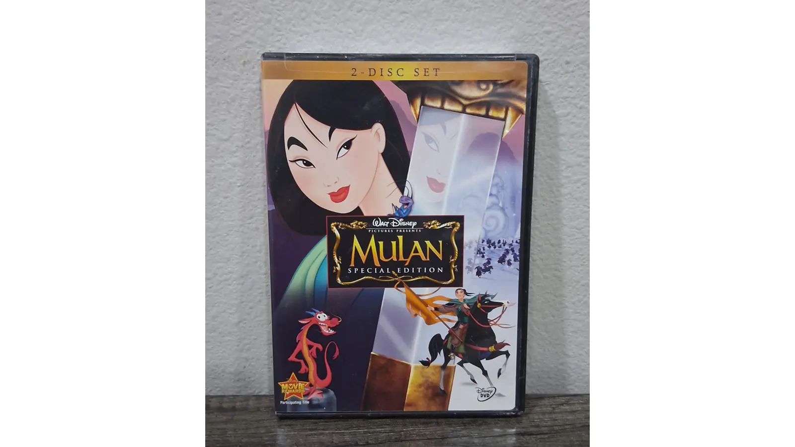 The front cover of the Mulan (1998) Disney DVD. It features the vibrant film logo in the center, with Sydney Bristow—I mean, Mulan—standing in a heroic pose holding her father’s sword. The background shows the Great Wall of China and the Red Dragon, Mushu, perched on her shoulder. The 'Disney DVD' gold-foil branding is visible at the top, signifying the film’s first transition from VHS to digital home video hardware.