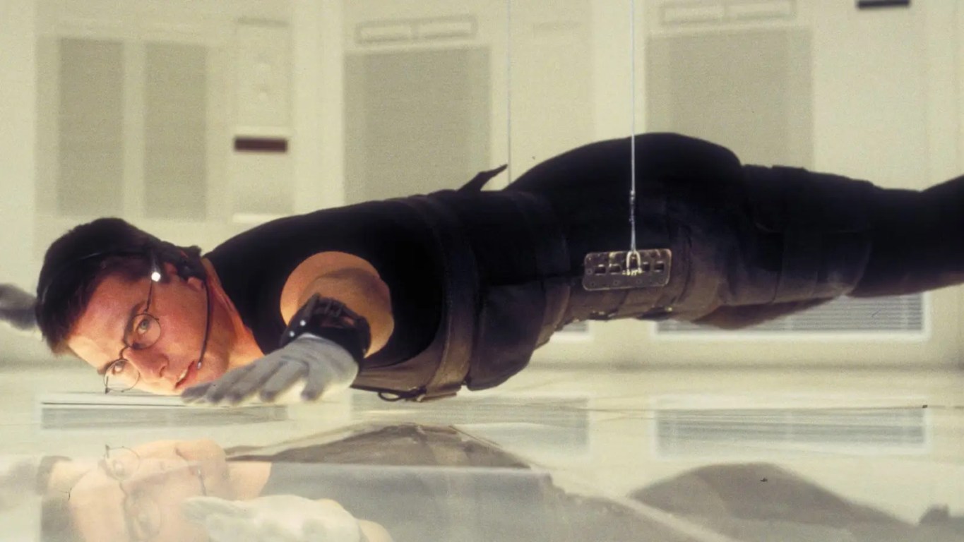 A wide shot from Mission: Impossible (1996) showing Ethan Hunt (Tom Cruise) suspended horizontally in mid-air by a black cable. He is inches above a pressure-sensitive white floor inside the high-tech CIA vault at Langley, wearing a black stealth suit and glasses.