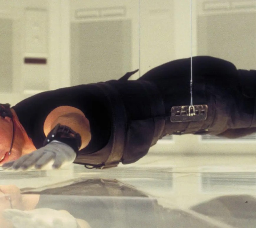 A wide shot from Mission: Impossible (1996) showing Ethan Hunt (Tom Cruise) suspended horizontally in mid-air by a black cable. He is inches above a pressure-sensitive white floor inside the high-tech CIA vault at Langley, wearing a black stealth suit and glasses.