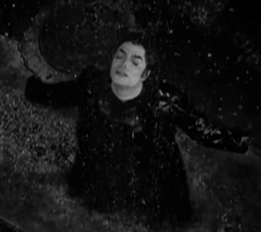 Black‑and‑white overhead shot of Michael Jackson standing alone in the rain with arms outstretched, head tilted back, and eyes closed, capturing the isolation and emotional intensity of the ‘Stranger in Moscow’ short film.