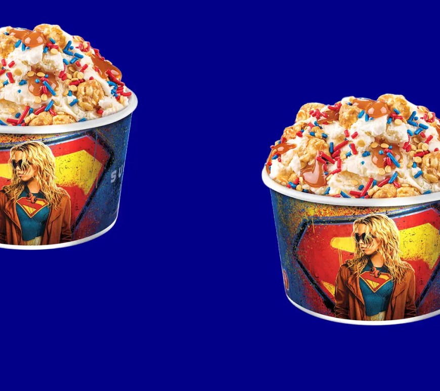 A crisp studio profile of Cold Stone's Supergirl Cosmic Kettle Corn Signature Creation in a branded cup, layered with golden caramel corn and red and blue star sprinkles.