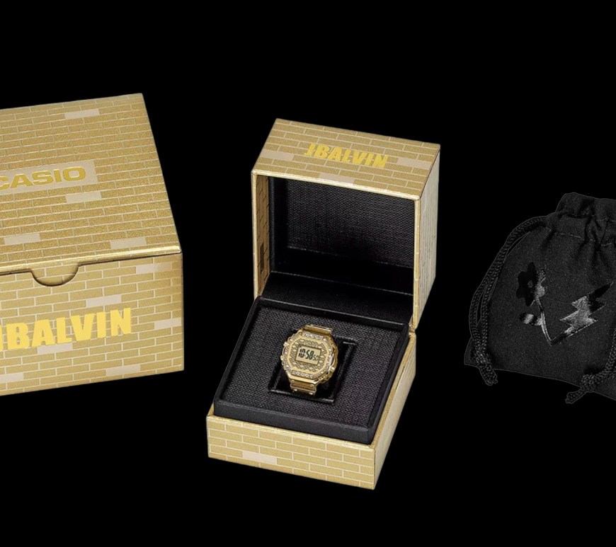 Studio product shot of the gold-tone Casio x J Balvin digital ring watch, showcasing its miniature LCD screen, embossed gold-brick texture, and rhinestone-encrusted bezel.