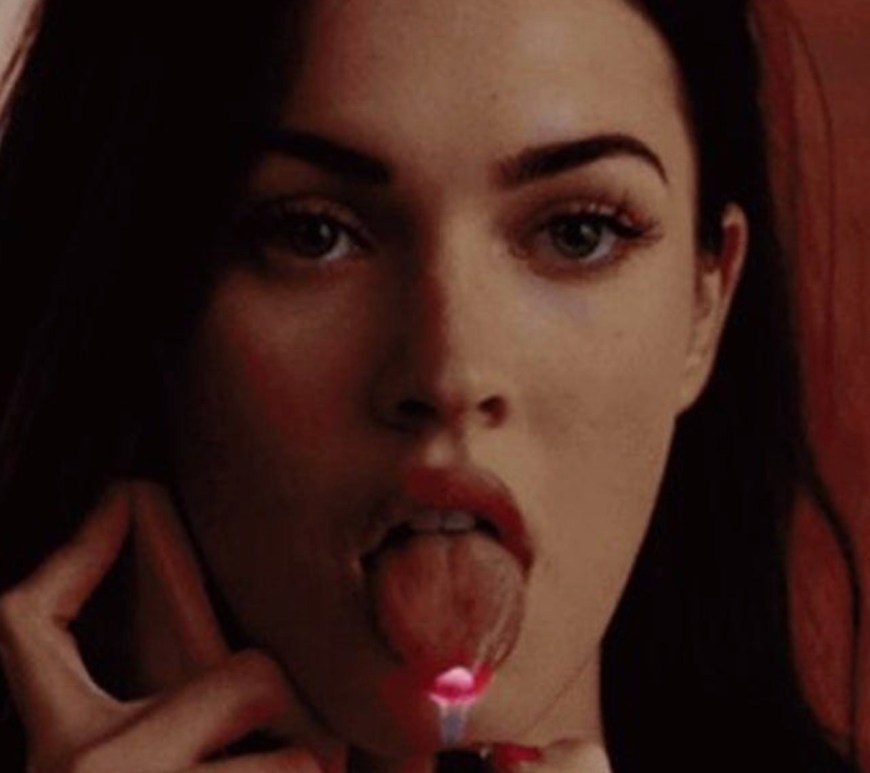 Close-up cinematic still of Megan Fox in Jennifer's Body holding a lit silver lighter directly to her tongue, showcasing the famous supernatural horror scene.