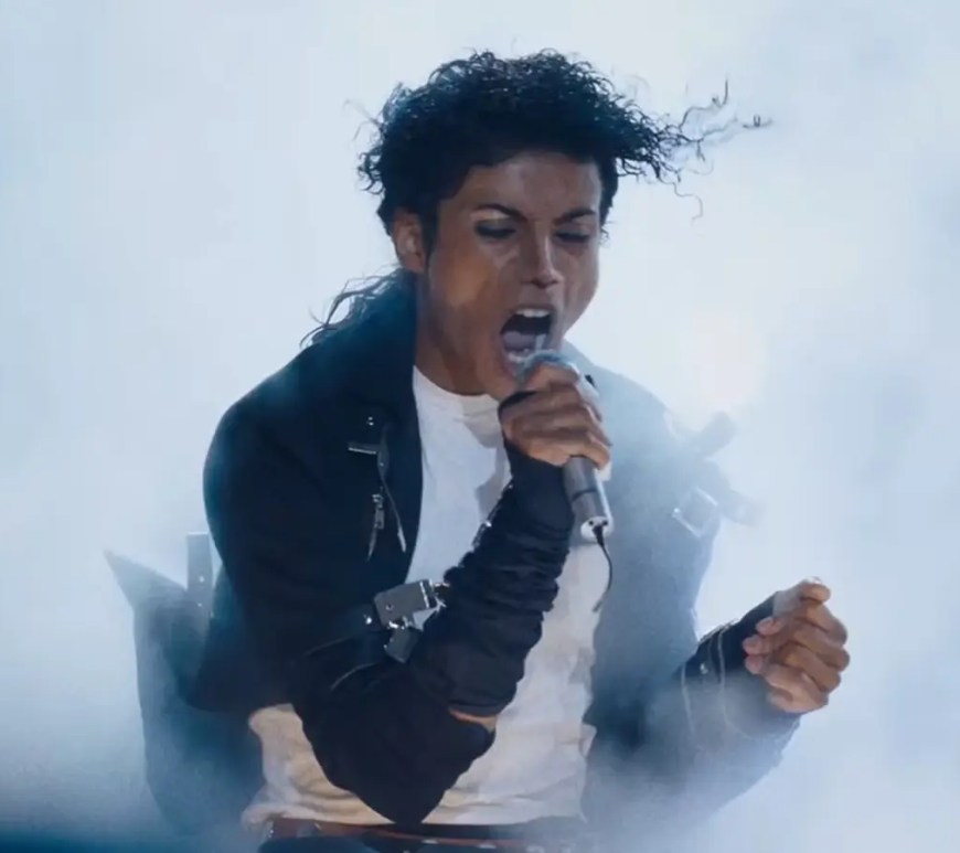 A theatrical production still from the 2026 film MICHAEL, featuring Jaafar Jackson performing as Michael Jackson on stage into a microphone, surrounded by cinematic stage smoke, dramatic spotlighting, and a high-contrast dark backdrop.