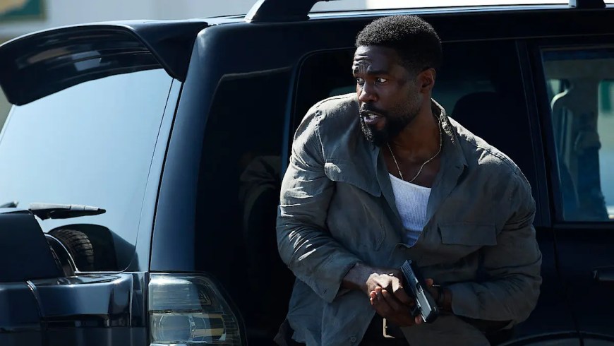 A close-up promotional shot of Yahya Abdul-Mateen II as John Creasy in the 2026 Netflix series Man on Fire. He is depicted with a focused, intense expression, his face partially shadowed to highlight the gritty, atmospheric tone of the show. He is dressed in tactical, lived-in gear suitable for the Rio de Janeiro setting. In the background, the soft-focus lights of a Brazilian favela at dusk create a high-contrast, cinematic feel, emphasizing his role as a lethal yet haunted protector.