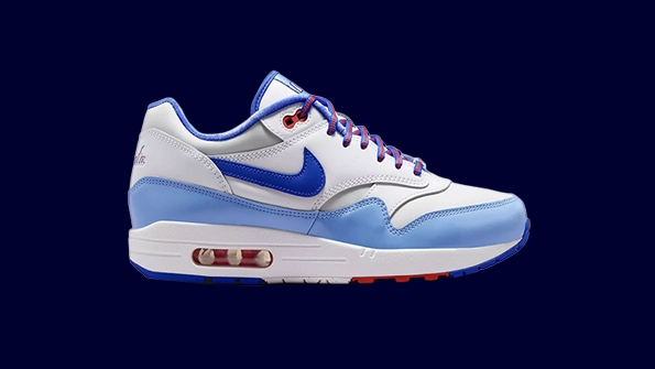 A side profile shot of the Nike Air Max 1 “Atlanta Braves” City Connect sneaker. The design features a crisp white mesh and leather base with a layered mudguard in light powder blue and deep royal blue, inspired by the Braves' 1974 throwback uniforms. A bold navy blue Swoosh sits on the mid-panel, complemented by university red accents on the upper eyelets and heel branding. The classic white midsole houses a visible Air Max unit, while the heel displays a custom red "Atlanta" script logo, capturing the fusion of MLB heritage and modern streetwear against a blue background.