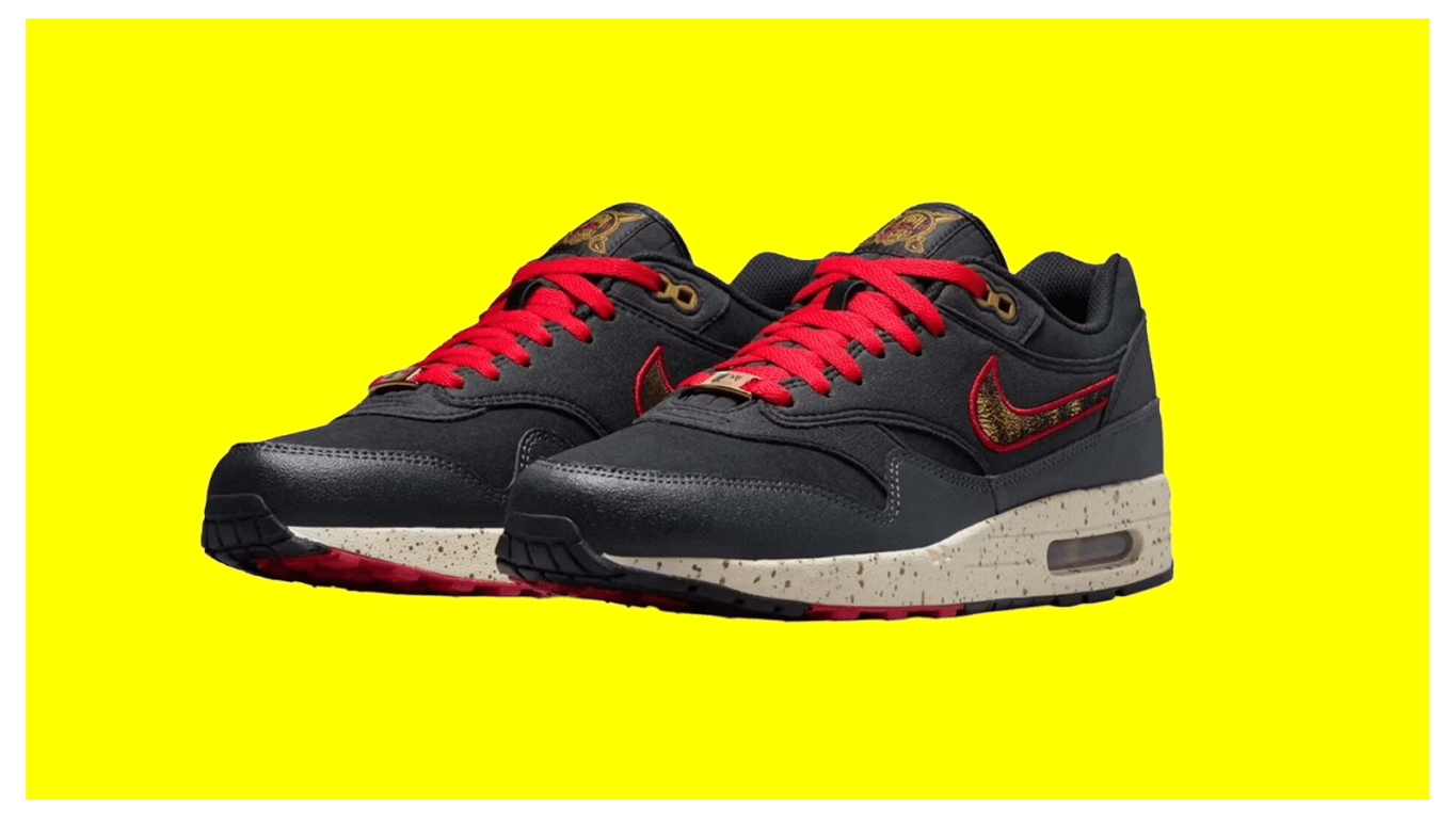 A side profile view of the Nike Air Max 1 City Connect "Pittsburgh Pirates" (SKU: IQ2893-001). The sneaker features a dark, tactical design with a black nylon canvas base and premium black nubuck leather overlays along the mudguard and midfoot. A prominent textured chenille Swoosh in Gym Red with black horizontal stripes—inspired by the "Jolly Roger" bandana—is positioned on the side panel. The shoe includes metallic gold top eyelets and red laces for high-contrast detail.