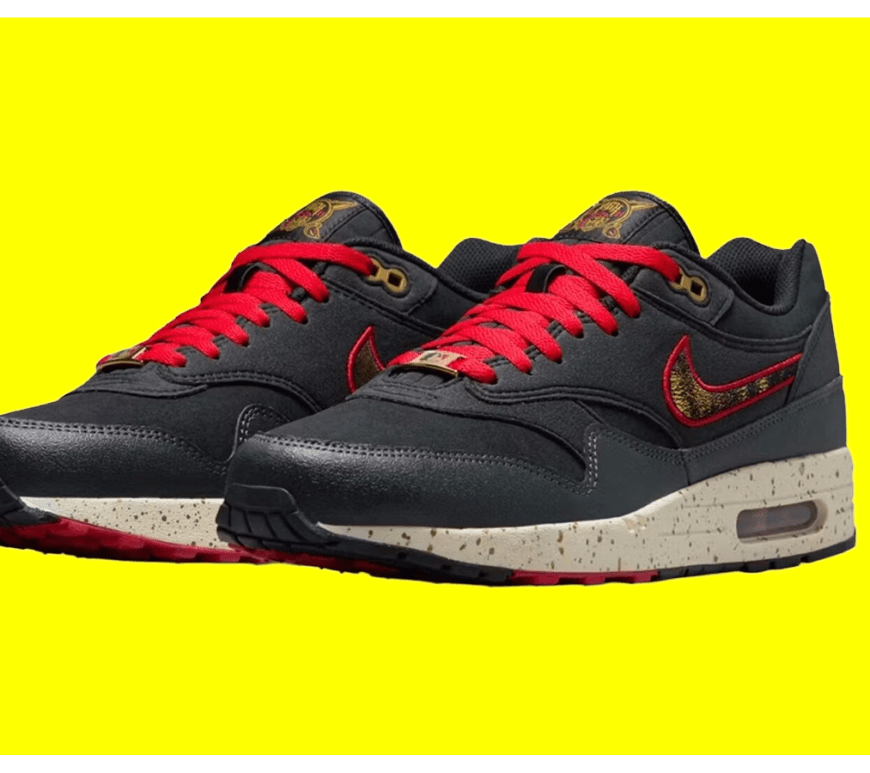 A side profile view of the Nike Air Max 1 City Connect "Pittsburgh Pirates" (SKU: IQ2893-001). The sneaker features a dark, tactical design with a black nylon canvas base and premium black nubuck leather overlays along the mudguard and midfoot. A prominent textured chenille Swoosh in Gym Red with black horizontal stripes—inspired by the "Jolly Roger" bandana—is positioned on the side panel. The shoe includes metallic gold top eyelets and red laces for high-contrast detail.