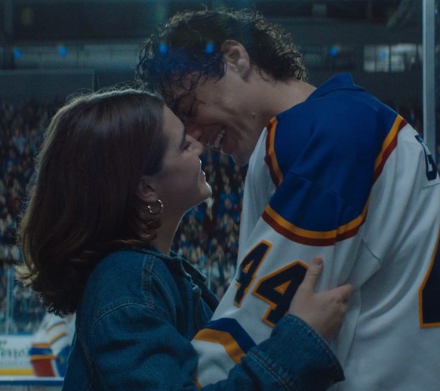 promotional still from Prime Video's Off Campus featuring the two main characters, Hannah Wells (Ella Bright) and Garrett Graham (Belmont Cameli). Hannah, a music student with long, wavy brown hair, stands on the left wearing a casual knit sweater and looking up with a clever, slight smile. Garrett, the Briar University hockey captain, stands close to her on the right wearing his blue and gold varsity hockey jacket over a gray hoodie, looking down at her with an intense, focused expression that hints at their onscreen chemistry.