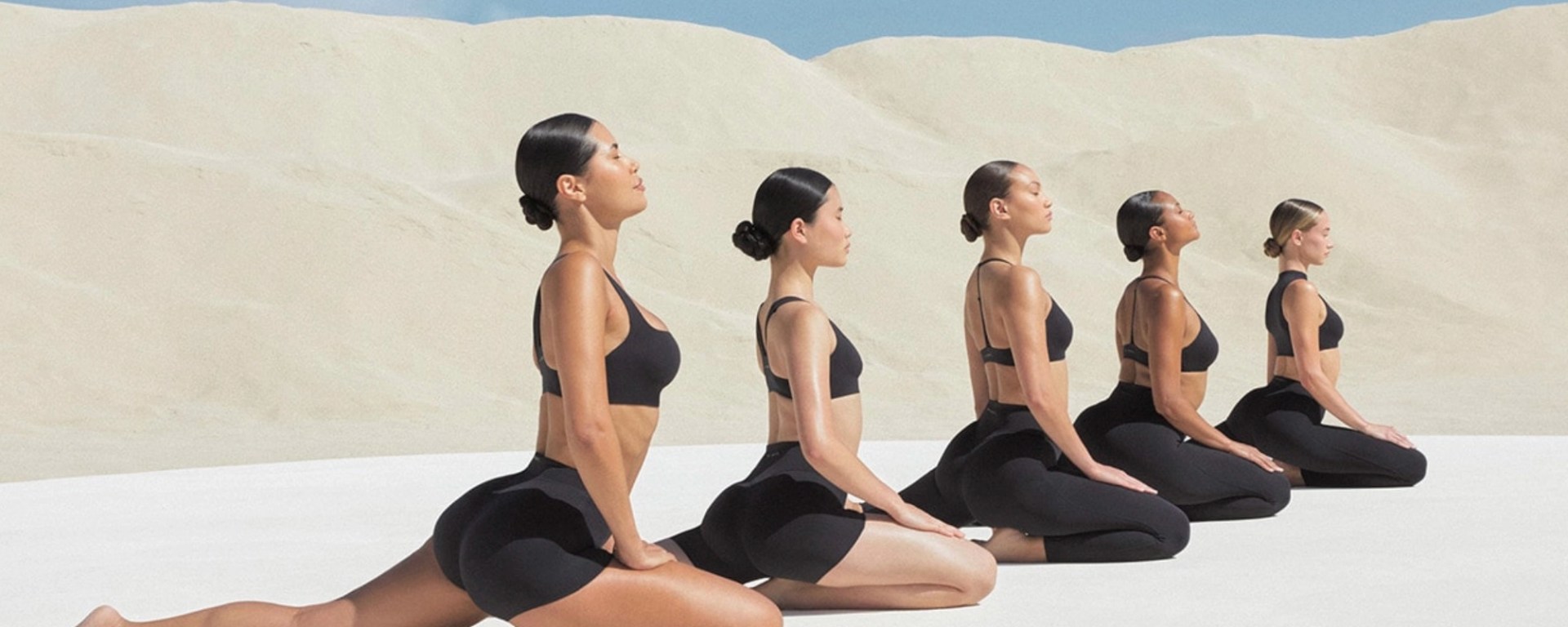 A high-resolution campaign image of a model performing a yoga pose in a minimalist studio. She is wearing the NikeSKIMS Studio Stretch obsidian leggings and a matching sports bra. The lighting is soft and neutral, highlighting the smooth, second-skin texture of the buttery fabric and the seamless design of the activewear.
