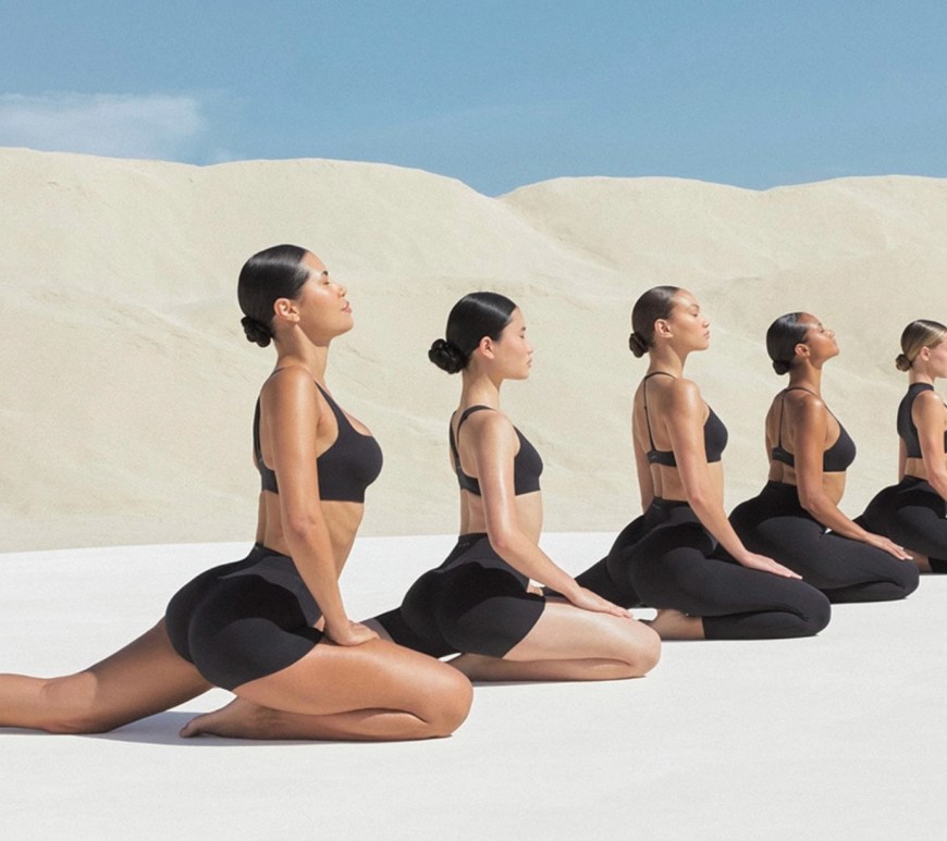 A high-resolution campaign image of a model performing a yoga pose in a minimalist studio. She is wearing the NikeSKIMS Studio Stretch obsidian leggings and a matching sports bra. The lighting is soft and neutral, highlighting the smooth, second-skin texture of the buttery fabric and the seamless design of the activewear.