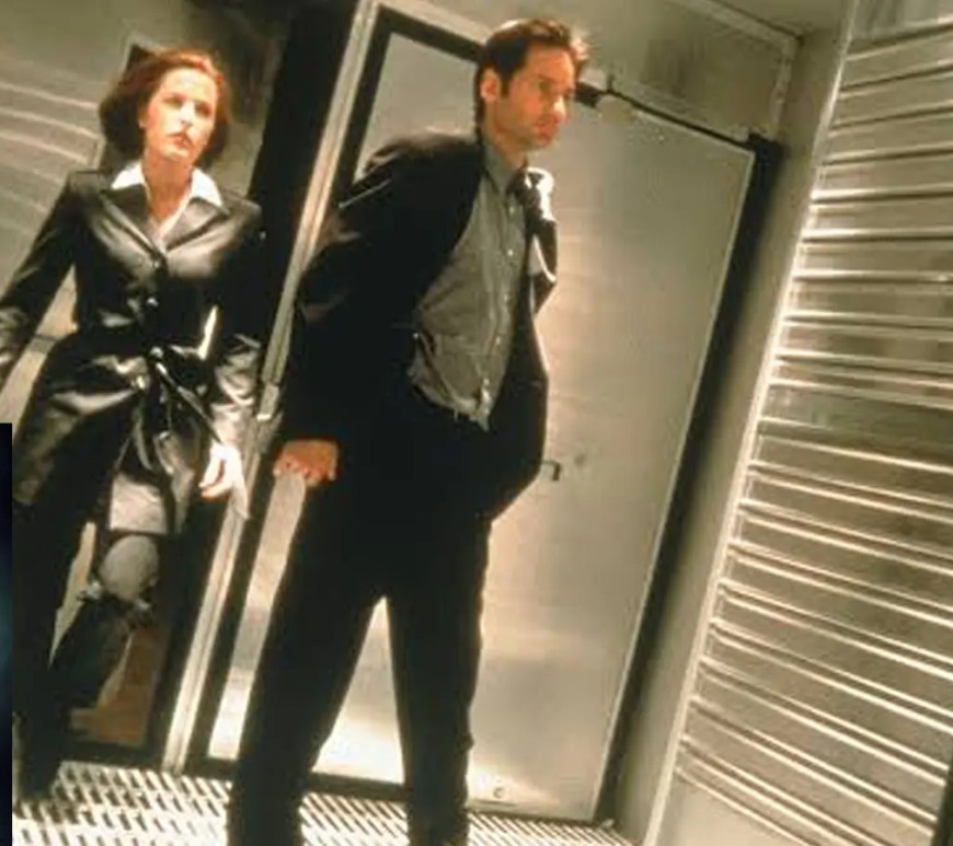 A tense, high-contrast cinematic film still from the 1998 movie The X-Files, showing David Duchovny as Fox Mulder and Gillian Anderson as Dana Scully investigating a dark corridor with flashlights.